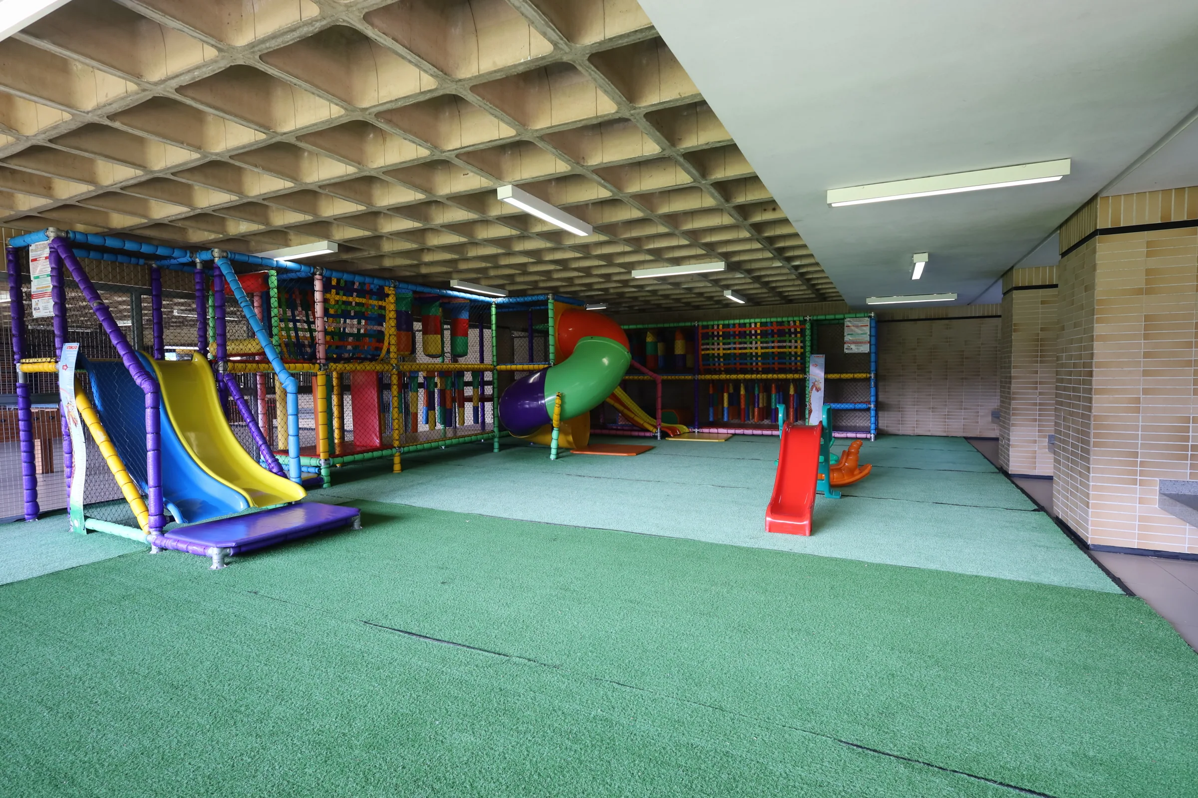 Vista ampla do Espaço Kids com brinquedo, escorregadores e piso verde