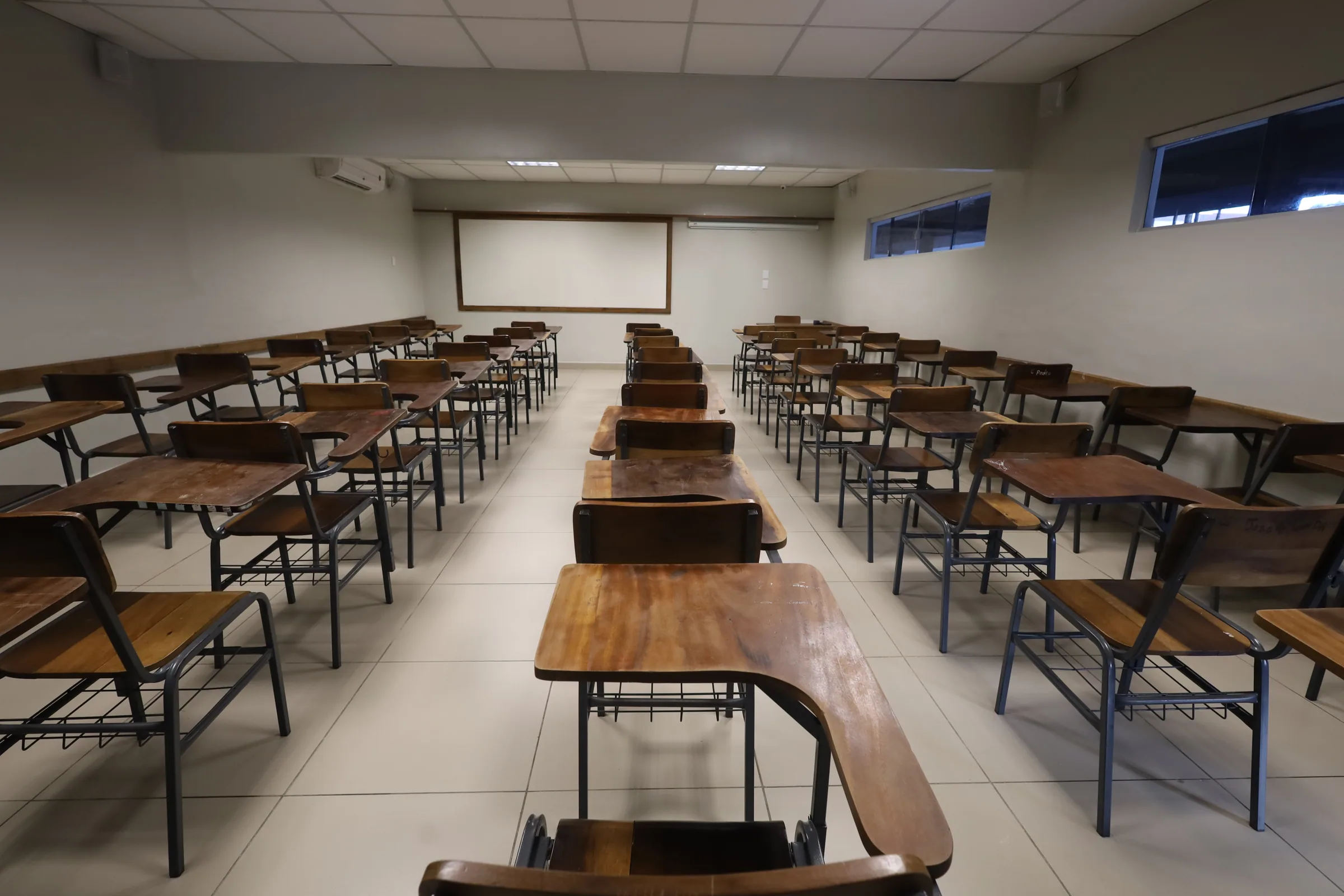 Vista do fundo de uma sala de aula mostrando carteiras, quadro e janelas